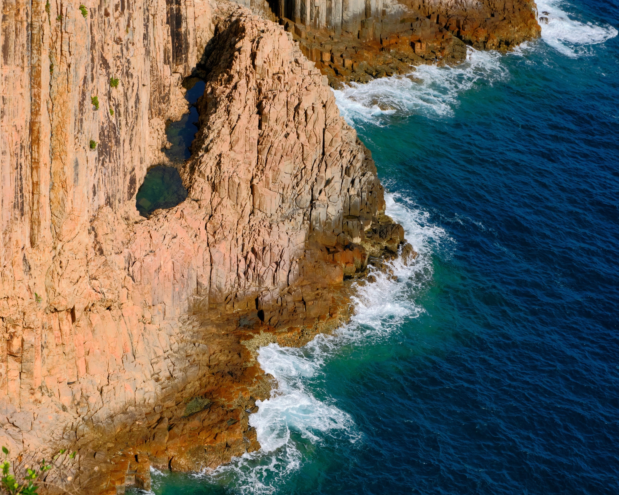 Orange-brown rock cliff meets deep blue ocean with white surf, showing a small cave entrance