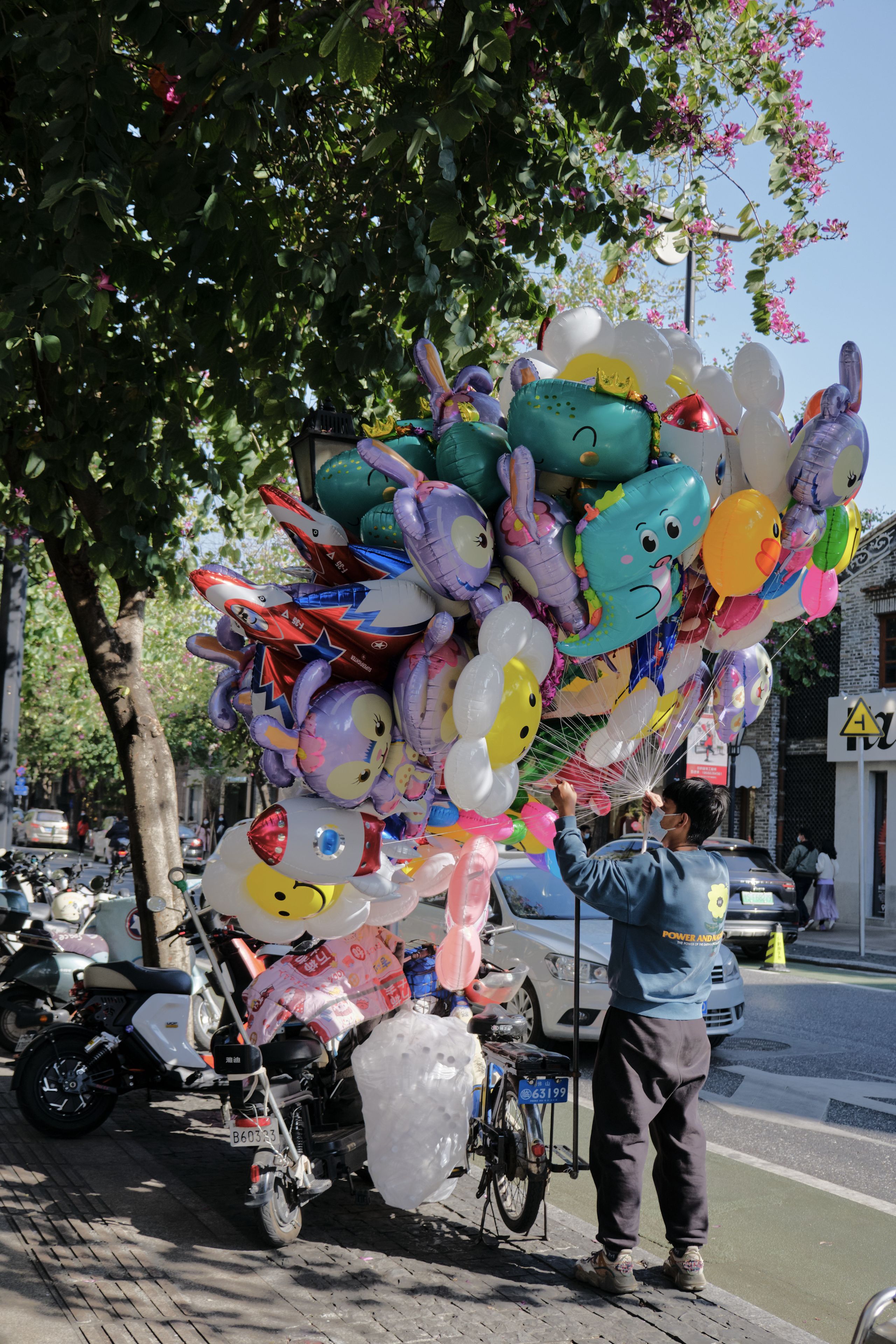 A person stands next to a large cluster of multicolored, character-shaped balloons attached to a cart on a sunny outdoor street. Parked scooters and trees are visible in the background