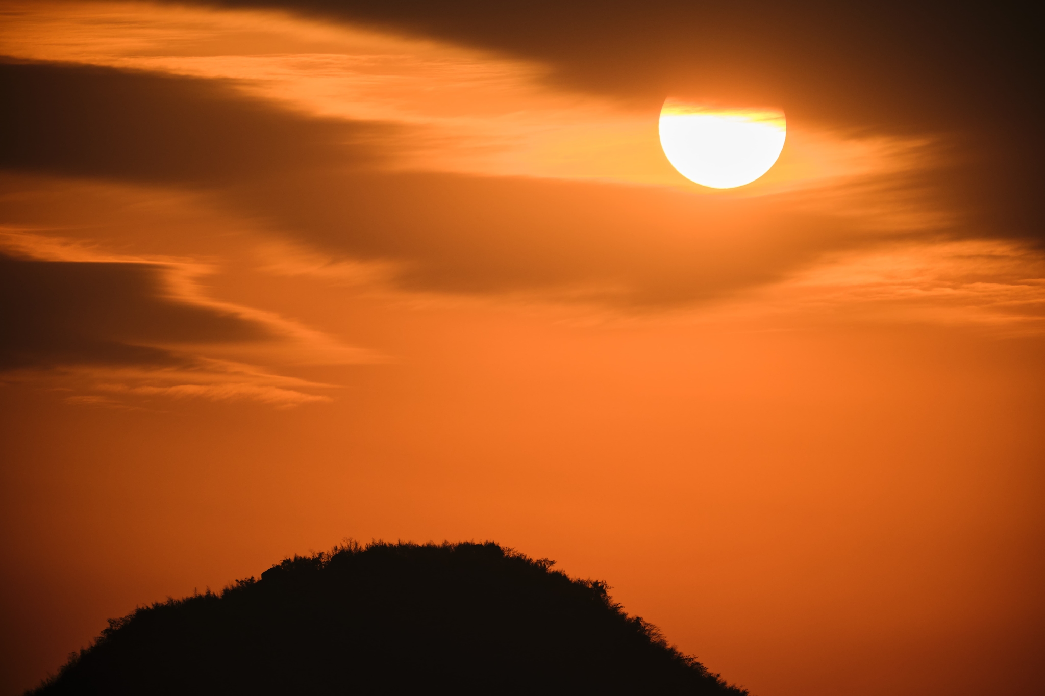 Half sun visible in an orange sky with clouds above a dark silhouetted hill