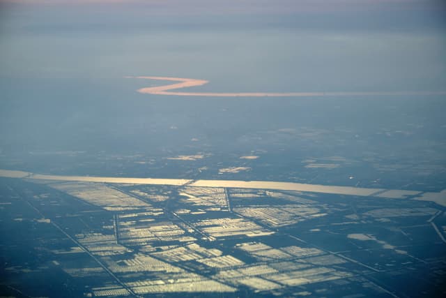 Aerial view of a reddish, winding river flowing through a hazy, snow-dusted landscape featuring rectangular fields and a wider river