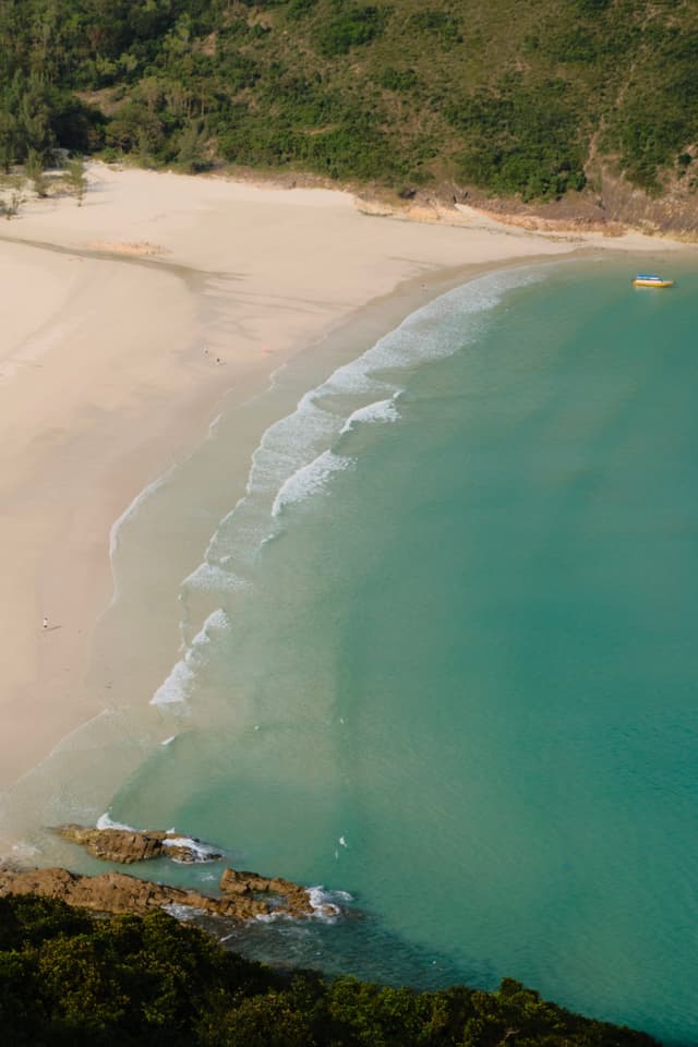 Aerial view of a crescent white sand beach meeting turquoise ocean water with gentle waves, framed by green hills and rocky coastline