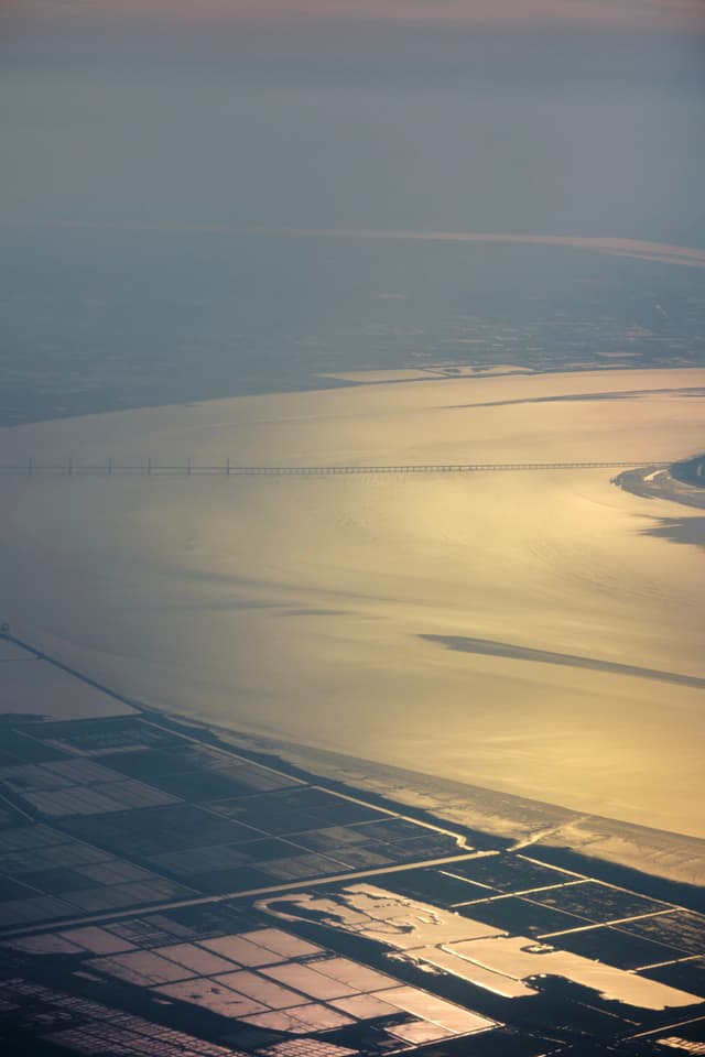 Aerial view of a wide, sunlit estuary bordered by geometric fields and distant land, with a faint bridge across