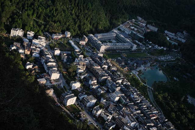 Aerial view of a village with numerous buildings bordered by a dark green forest, featuring a small body of water