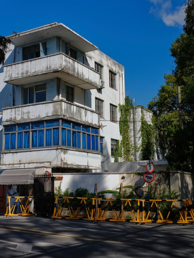White three-story building with multiple balconies and prominent blue windows on the second floor, bordered by a white wall and numerous yellow barriers on a dark street, under a clear blue sky