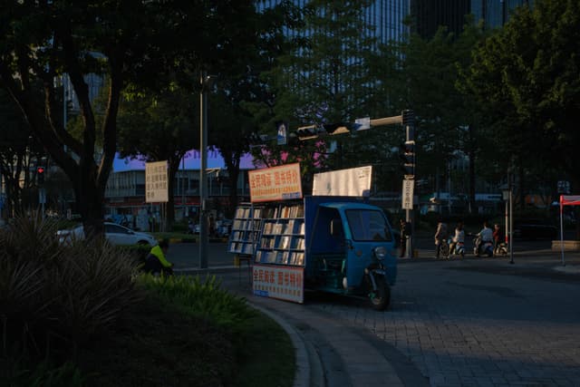 A blue three-wheeled vehicle modified into a mobile bookstore drives on a shaded urban street. Trees line the background, with distant buildings and pedestrians