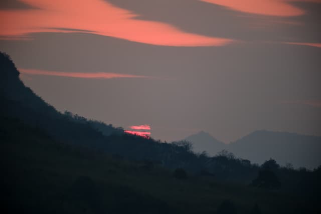 Red sun partially visible behind a dark mountain range, with silhouetted hills and reddish wavy clouds in a grey sky