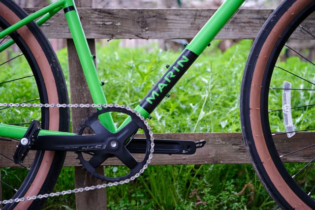 Close-up of a bright green bicycle frame, crankset, chain, pedals, and parts of wheels with brown sidewalls against a blurred background of green grass and a wooden fence
