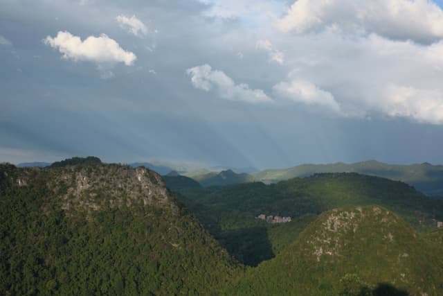 Forested mountain range and valley under a cloudy sky with sun rays and a faint rainbow