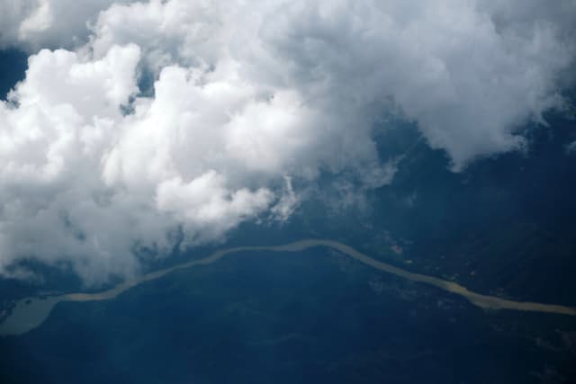 Aerial view of a winding river through a dark, forested landscape, partially obscured by white cumulus clouds