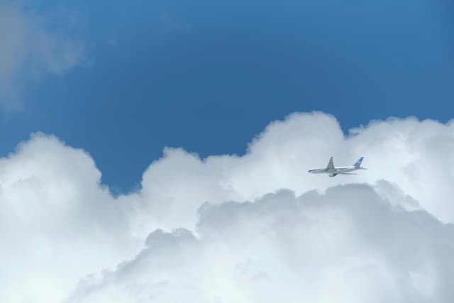 A white passenger airplane flies horizontally above a layer of fluffy white clouds against a clear blue sky