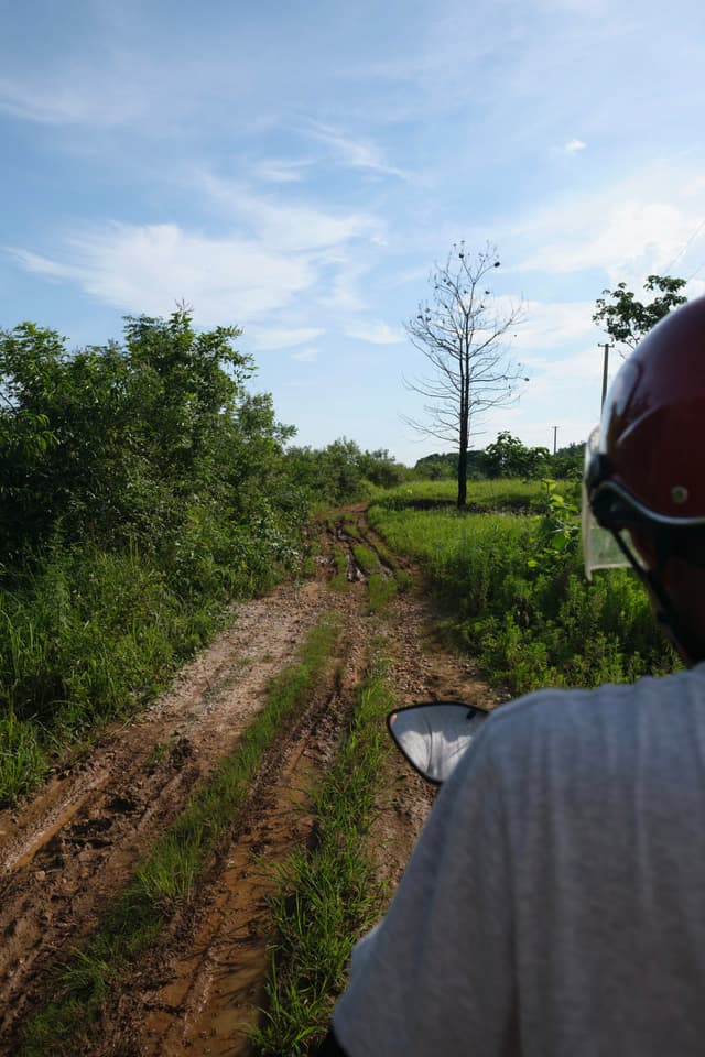 View from behind a person wearing a red helmet on a muddy dirt path with grass and trees under a partly cloudy sky