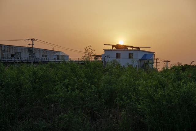 Sunset over industrial-style buildings and dense green foliage under a hazy sky with power lines visible