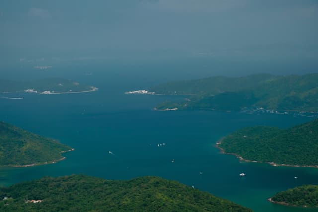 Aerial view of a blue-green sea inlet flanked by lush green, forested islands with multiple white boats, under a hazy sky