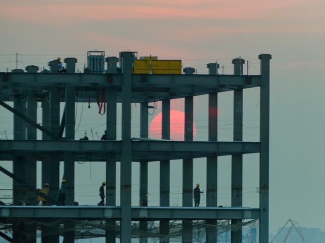 Steel framework of a multi-story building under construction with a large red sun visible through its upper levels. Silhouetted workers are on the lower floors against a pale sky