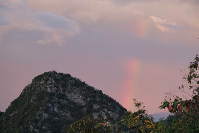 Dark forested mountain peak under a light pink sky with a faint vertical rainbow effect and sparse clouds, with foliage visible on the right