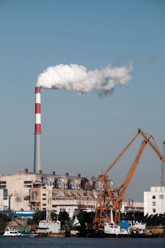 Smokestack emitting white smoke above an industrial building, with orange cranes and water in the foreground under a blue sky