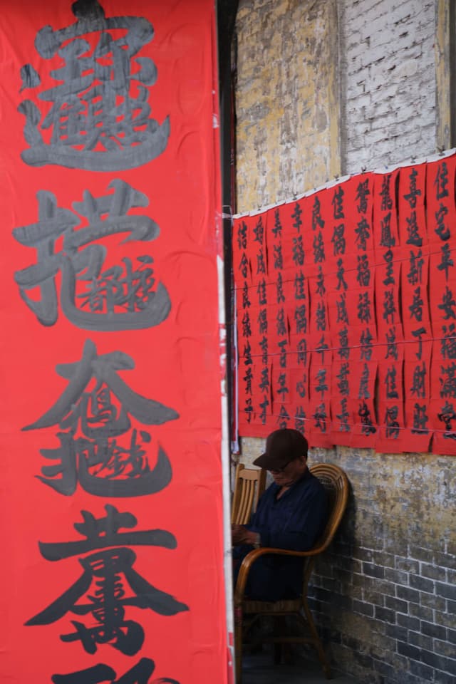A man sits on a wooden chair against a brick wall, partially obscured by a large red banner with black Chinese calligraphy. Smaller red banners with calligraphy hang on the wall to the right