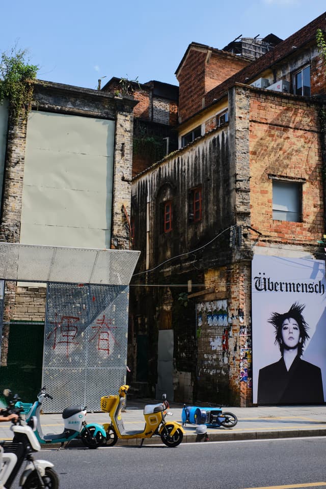 Weathered brick buildings and narrow passage, large 'Übermensch' billboard with a person, multiple scooters parked on urban street