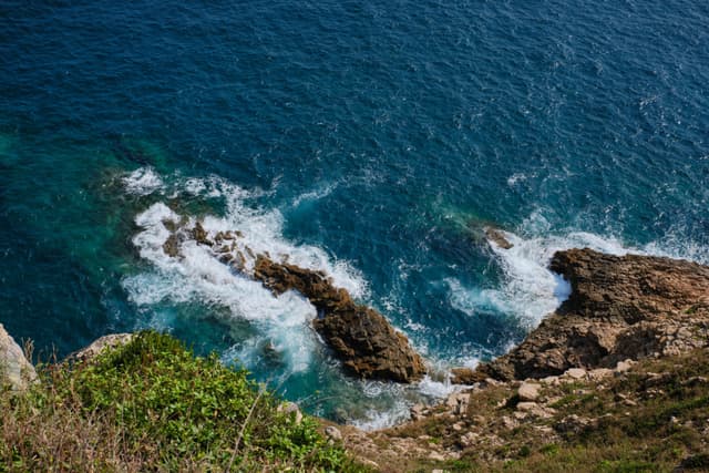 Deep blue ocean water with white waves crashing onto dark brown coastal rocks, seen from an elevated perspective with green vegetation at the edges