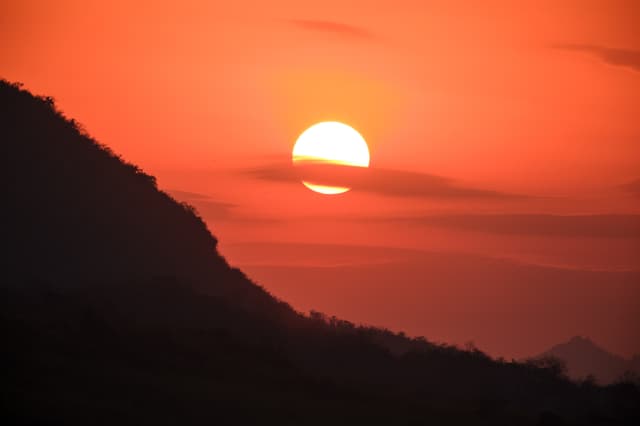 Bright sun centered against an orange and red sky, with a dark silhouetted mountain slope occupying the left foreground and subtle horizontal cloud bands