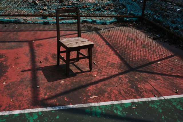 A single, old wooden chair sits on a weathered red court with faded white lines and shadows from a fence