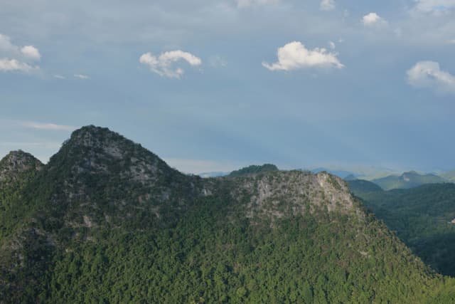 Dense green forests blanket rugged mountains under a blue sky with scattered white clouds