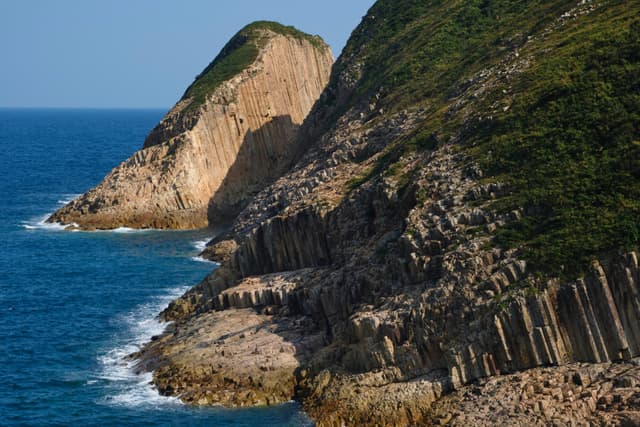 Hexagonal rock columns forming coastal cliffs alongside blue ocean with breaking waves and green vegetation under clear skies
