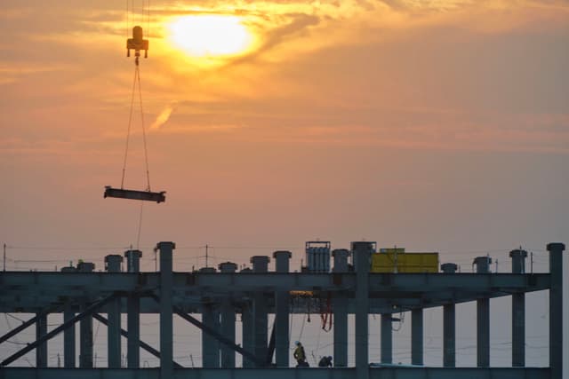 Orange sky with setting sun above silhouetted construction site. Crane lowers long object over concrete pillars and beams. Faint contrail present