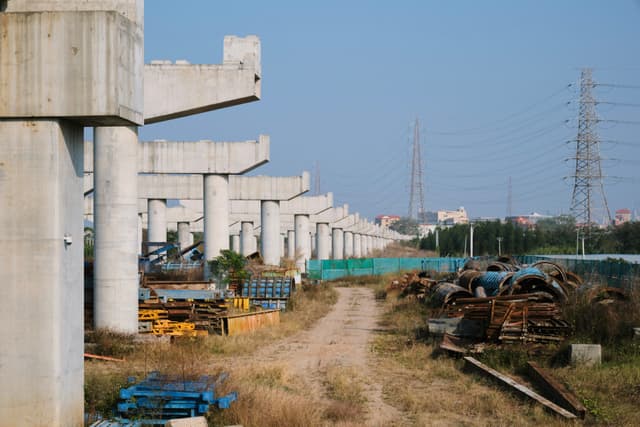 Row of concrete elevated infrastructure supports, dirt path, construction materials, power pylon, clear sky