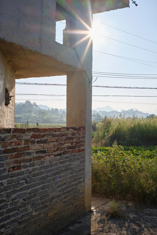 Sun flares from a concrete structure with an exposed brick wall, overlooking green fields and distant hills under a clear sky with power lines