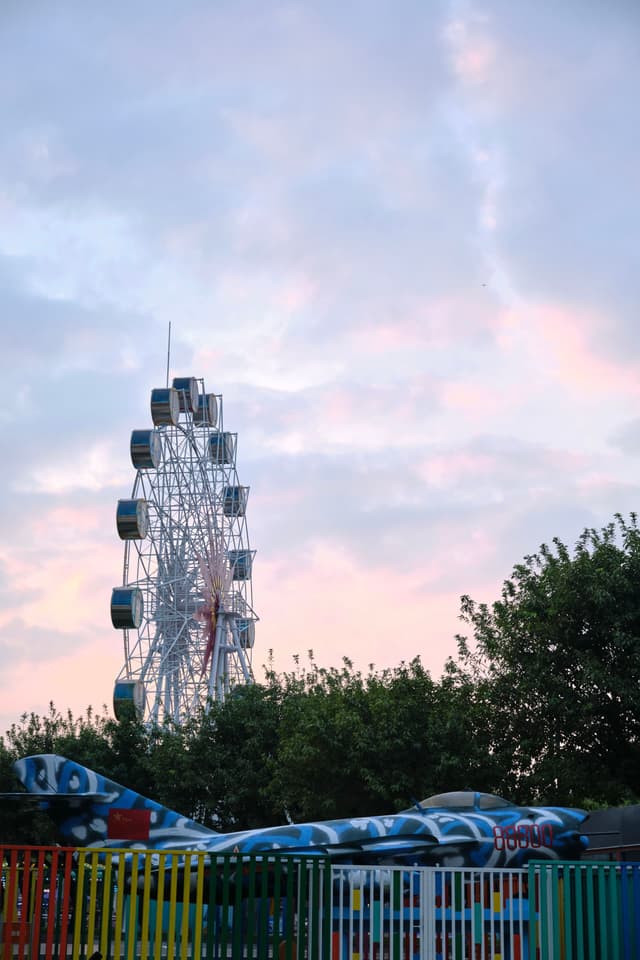 White Ferris wheel against cloudy pink-blue sky, airplane model, green trees, colorful fence