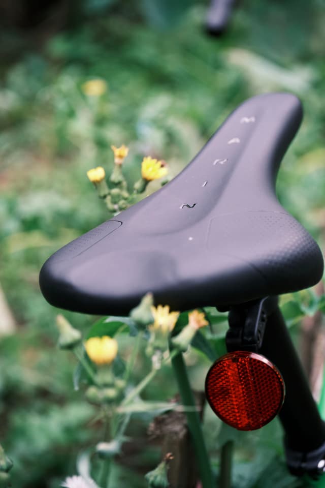 Black bicycle seat with water droplets and red reflector, against blurred green foliage and yellow flowers