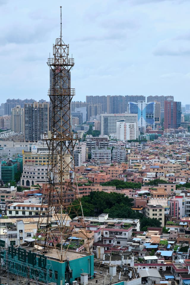Rusty lattice tower stands tall amid a dense urban landscape of low-rise buildings, with modern high-rises in the distant background under an overcast sky