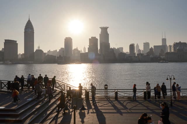 Sunlight glares over a city skyline across a wide river, silhouetting numerous people on a riverside promenade