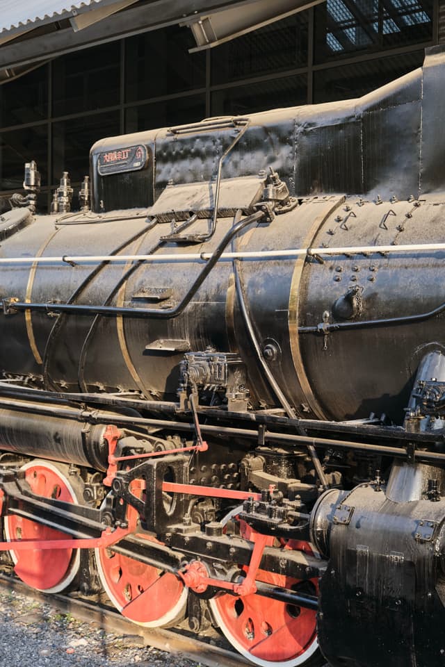 Black vintage steam locomotive with red wheels, gold banding, and intricate machinery, parked beneath a metal roof structure