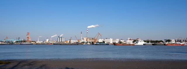 Panoramic view of an industrial port across a body of water with various buildings, cranes, and smokestacks under a clear blue sky. Smoke rises from several stacks. Several vessels are visible along the waterfront