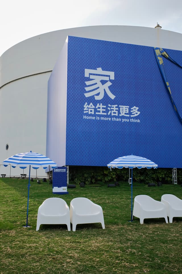 Grassy lawn with two white lounge chairs and two blue-and-white striped umbrellas in front of a building with a large blue banner displaying Chinese text and 'Home is more than you think'