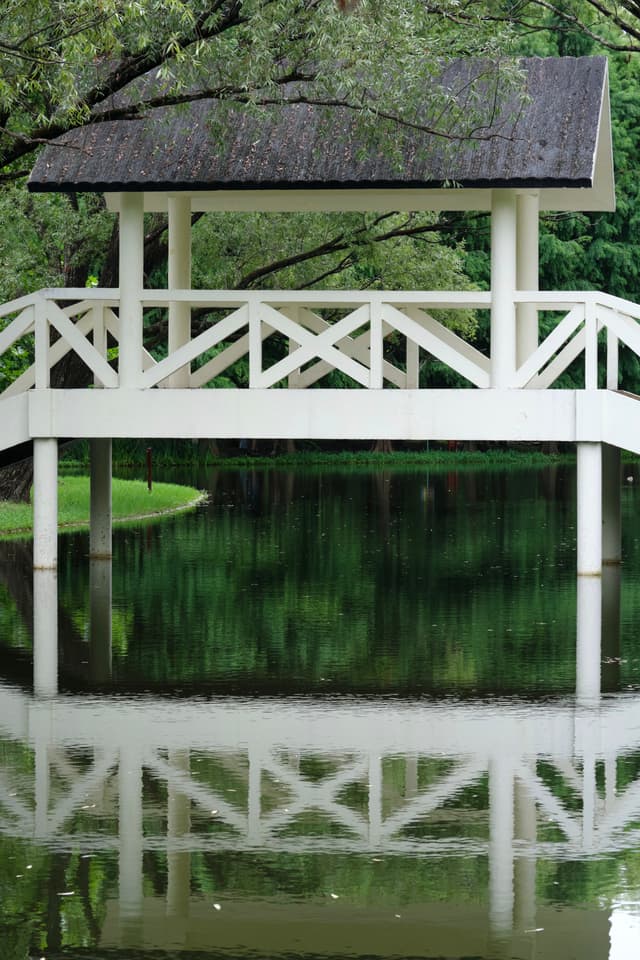 White gazebo with thatched roof stands over still water, reflecting its structure. Lush green foliage surrounds the scene