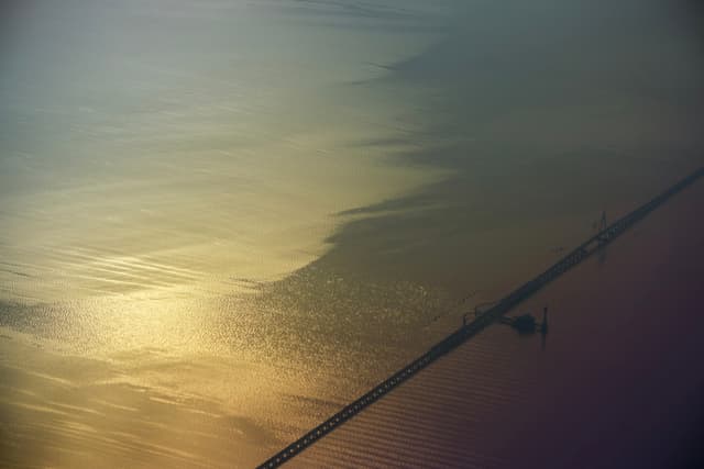 Aerial view of a long bridge over water, displaying a gradient of sunlit golden ripples to darker, hazy tones