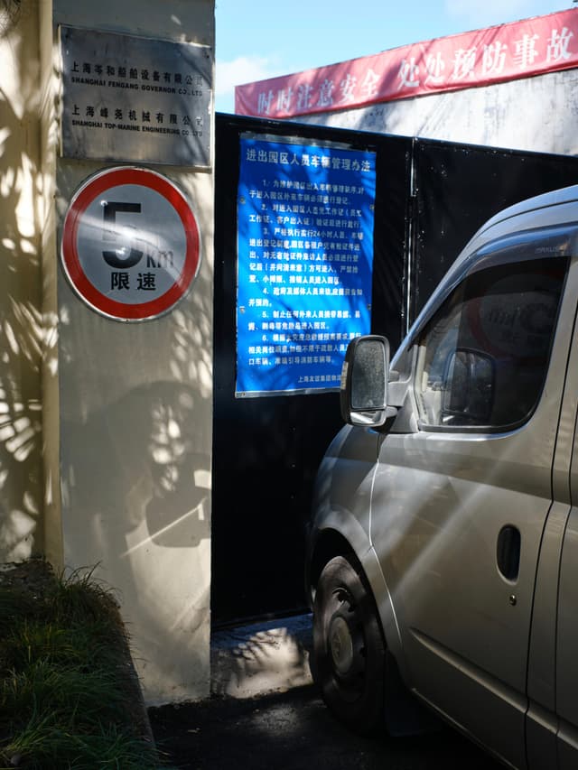 Silver van beside black gate, circular '5' speed limit sign, blue text sign, foliage shadows on wall