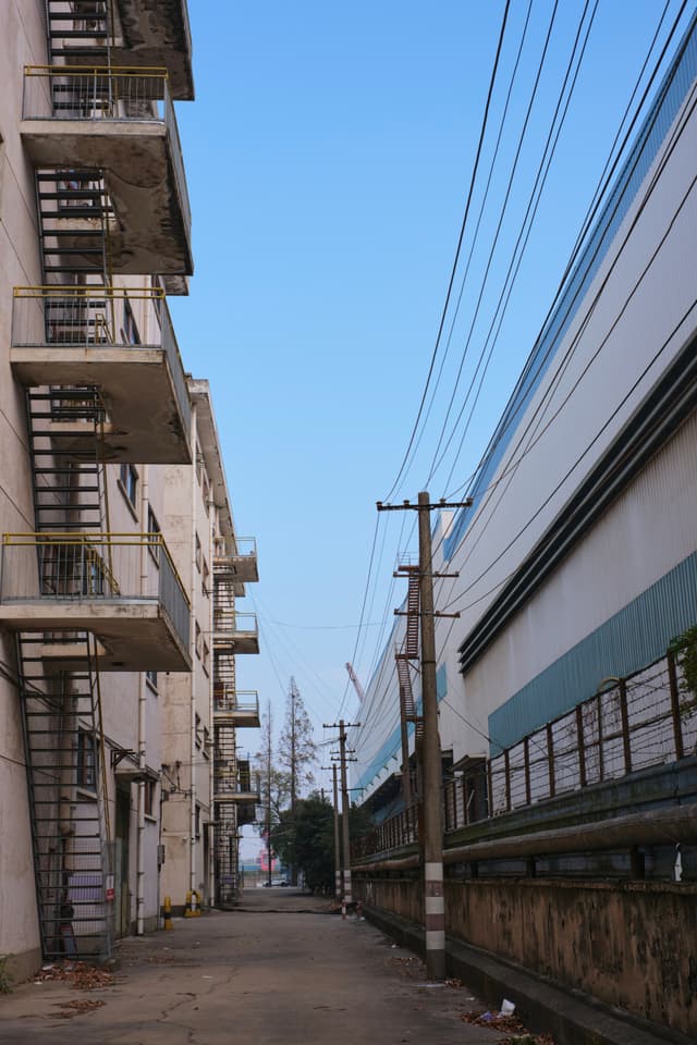 Narrow path between a multi-story building with external metal staircases and a plain-walled building, framed by utility poles and overhead power lines under a clear blue sky