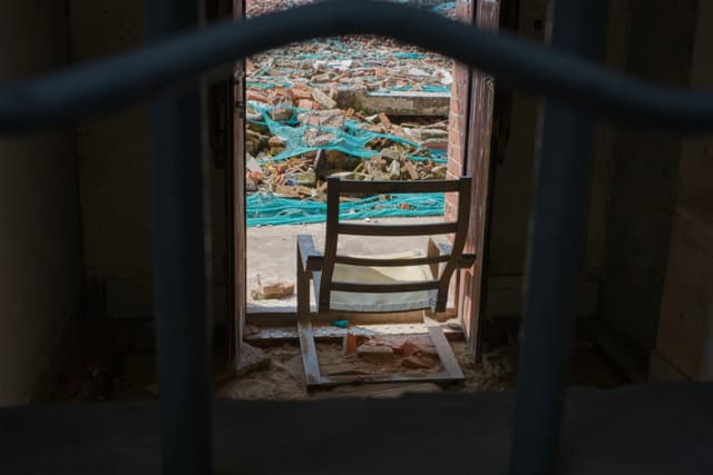Wooden chair in dark room facing open door to rocky, blue waters, viewed through blurred foreground bars