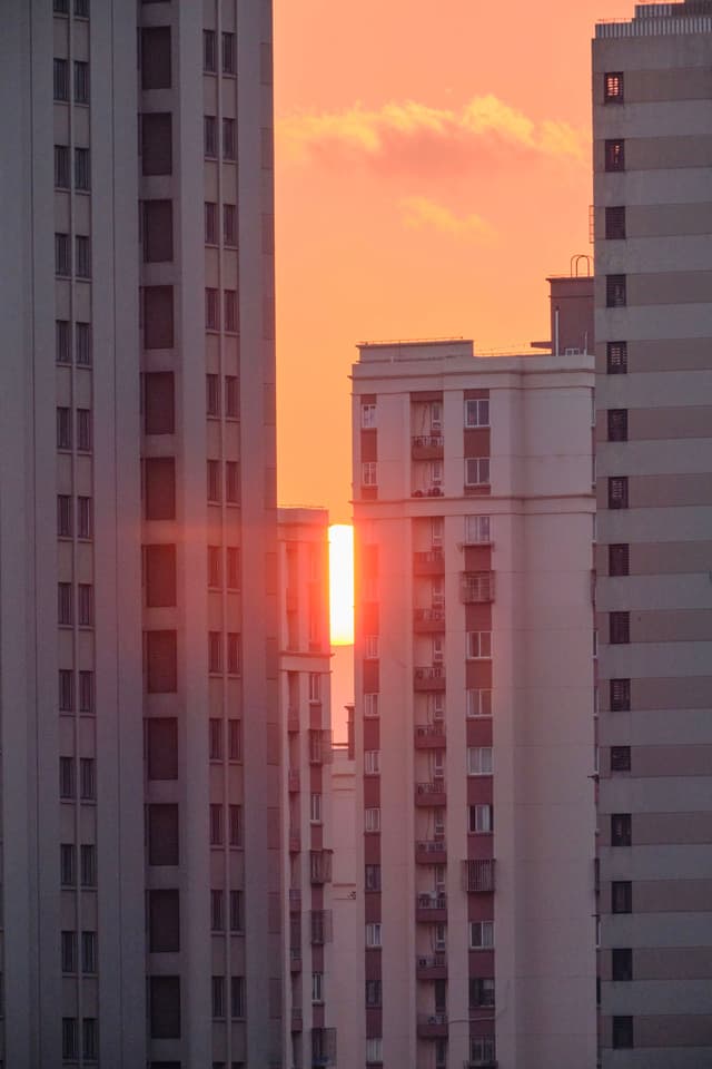 Vertical shot of orange sunset light slicing through gaps between tall, light-colored apartment buildings. Orange sky with clouds visible above