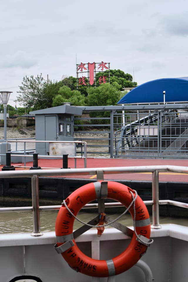 Orange lifebuoy on a white boat railing with a red-decked ferry terminal, gray building, blue canopy, and metal fence behind it. A large red sign with Chinese characters is visible on a distant building amidst green trees under an overcast sky