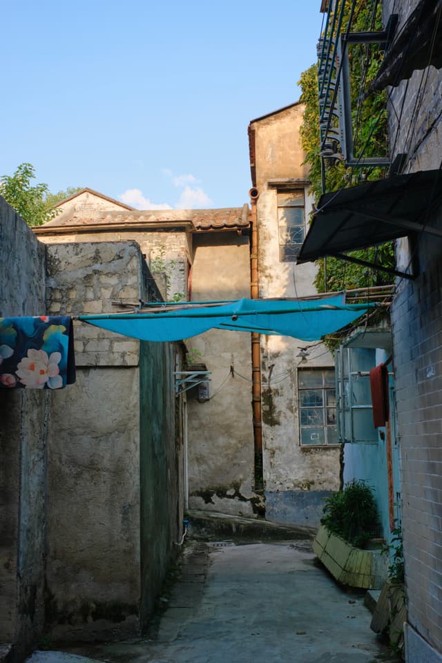Narrow outdoor alley between weathered buildings with laundry and blue tarp overhead