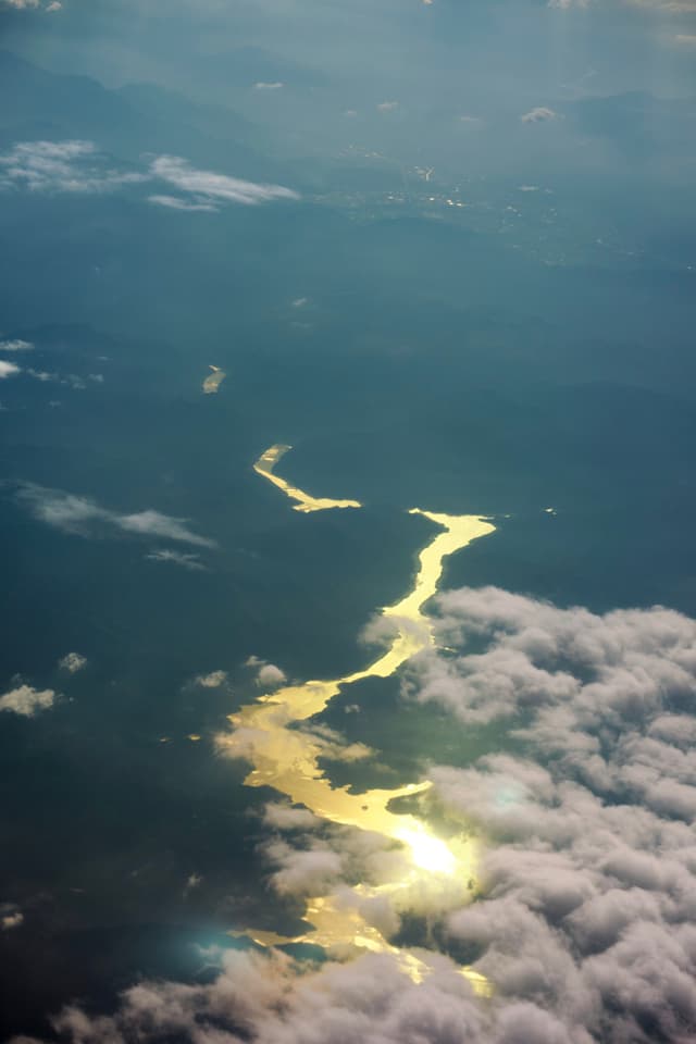 Aerial view of a winding, golden river reflecting sunlight, flowing through dark land partially covered by white and gray clouds