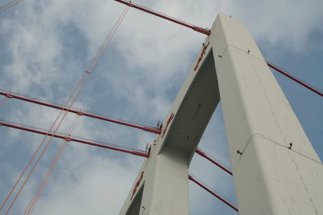 View looking up at concrete bridge towers and red suspension cables against a cloudy sky