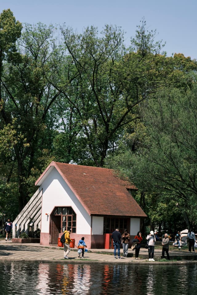 A white building with a red roof stands by a body of water. Several people are visible on the shore and near the building. Lush green trees and a clear sky form the background