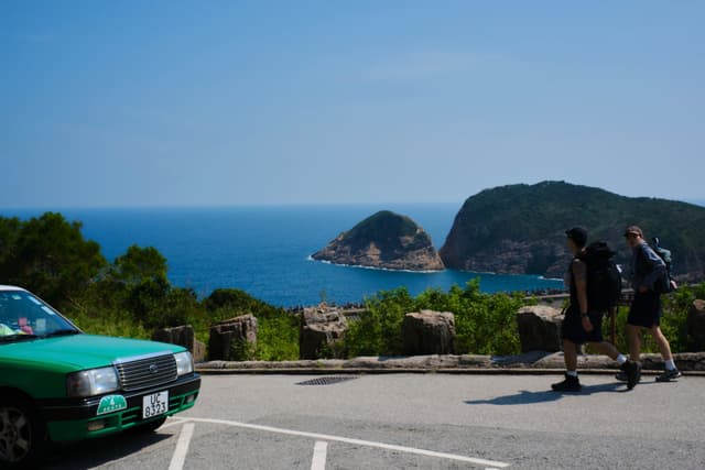 Green taxi, two pedestrians, blue ocean, distant islands, coastal road, clear sky