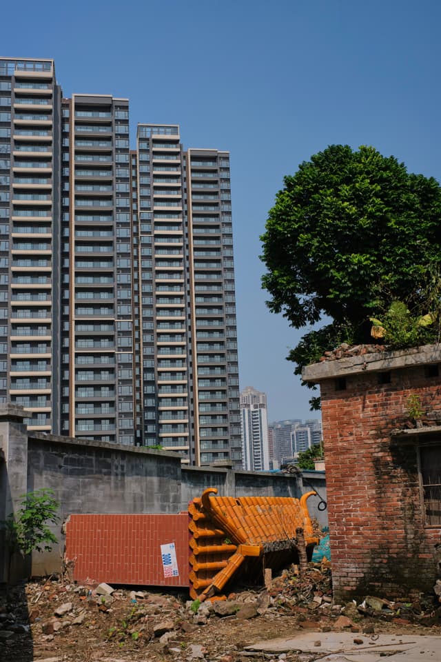 Modern high-rise buildings behind old brick structures, a collapsed orange tiled roof, and rubble under a clear blue sky